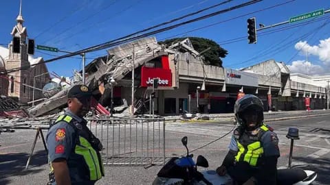 AFP via Getty Images Policemen in uniform stand in front of a barricaded road. Behind them is a partially collapsed building with a red Jollibee sign.